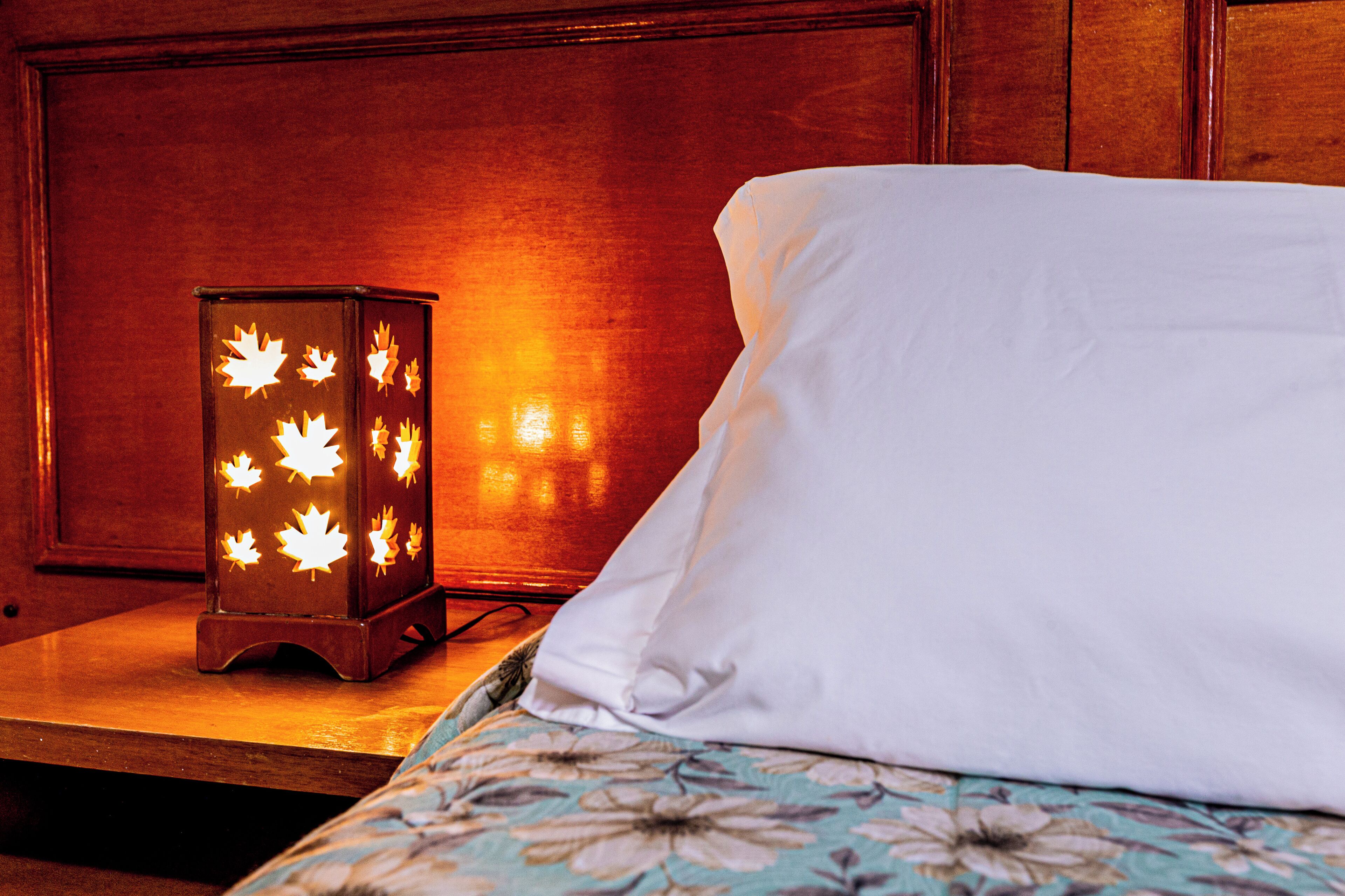 A bed with a white pillow and a floral-patterned blanket, a wooden nightstand with a maple-patterned lantern, and a wooden headboard.