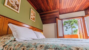 Une chambre avec un plafond en bois, un lit recouvert d’une literie à motifs floraux et une fenêtre donnant sur les arbres.