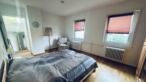 Un dormitorio con cama, una silla, una lámpara, un radiador y ventanas.
