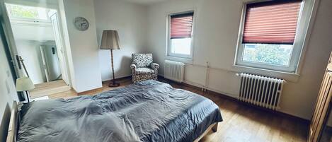 Ein Schlafzimmer mit Bett, Stuhl, Lampe, Heizkörper und Fenstern.