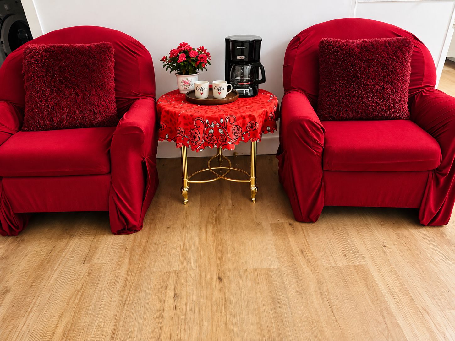 Deux fauteuils rouges ornés de coussins moelleux, une petite table avec une machine à café et un pot de fleurs, et une nappe rouge à motifs de roses.
