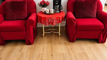 Deux fauteuils rouges ornés de coussins moelleux, une petite table avec une machine à café et un pot de fleurs, et une nappe rouge à motifs de roses.