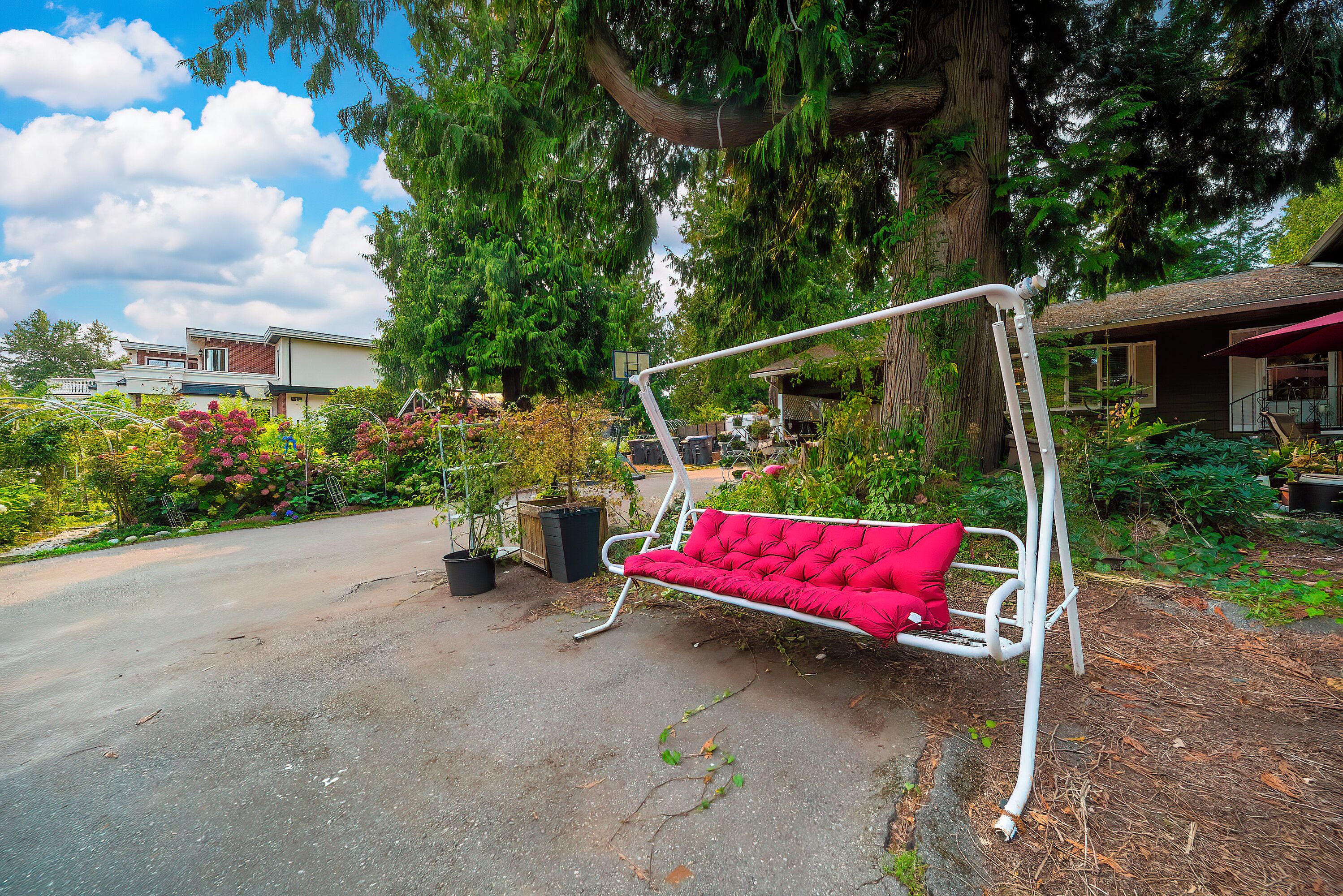 A red outdoor bench with a metal frame, surrounded by lush greenery and a large tree.