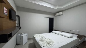 A hotel room with a bed, a TV, an air conditioner, and a small refrigerator.