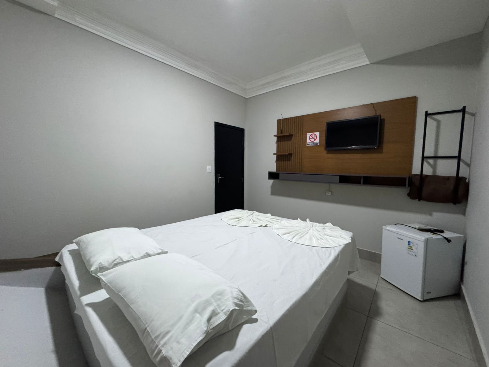A bedroom with a bed, a TV mounted on the wall, a small refrigerator, and a door.