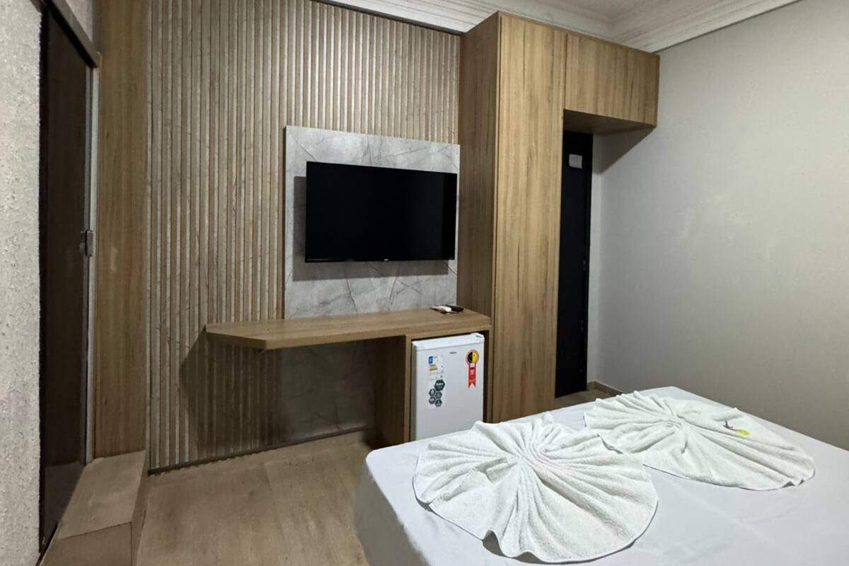 A hotel room with a TV mounted on a wooden panel wall, a small refrigerator, and a neatly made bed with towels.