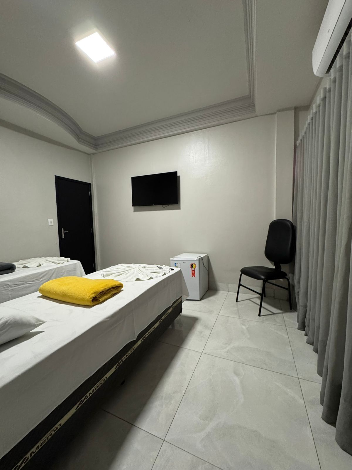 Quarto de hotel com cama, cadeira, televisão e um pequeno frigorífico.