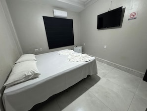 A single-bed room with a flat-screen TV, a black window blind, a wall-mounted air conditioner, and a no-smoking sign.