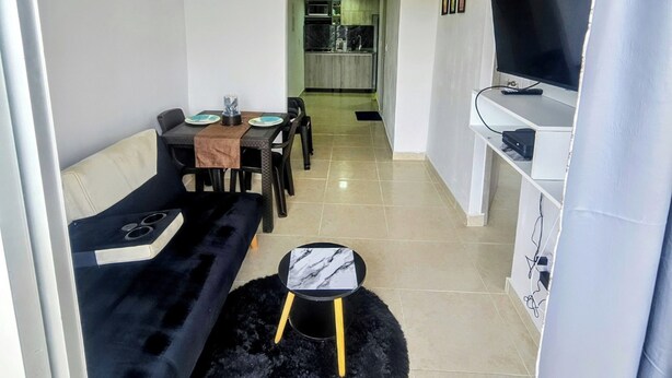 Un salon moderne avec un canapé noir, une petite table ronde et un téléviseur posé sur un meuble blanc. On aperçoit une cuisine au fond.
