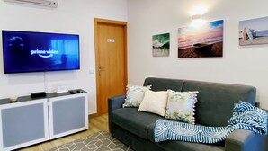 A living room with a grey sofa, floral cushions, a patterned rug, a flat-screen TV displaying 'prime video', and artwork on the wall.