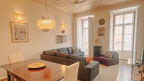 Un salón espacioso con una mesa de comedor de madera, un sofá gris y un sillón púrpura. Hay dos luces de pared y una araña.