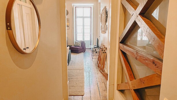 Un pasillo estrecho con vigas de madera, un espejo a la izquierda y una vista hacia una sala de estar con un sofá y un televisor.