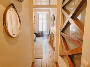 Un pasillo estrecho con vigas de madera, un espejo a la izquierda y una vista hacia una sala de estar con un sofá y un televisor.