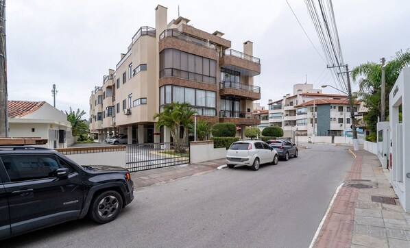 Um edifício residencial com vários andares, varandas, área de estacionamento murada e vista para a rua.