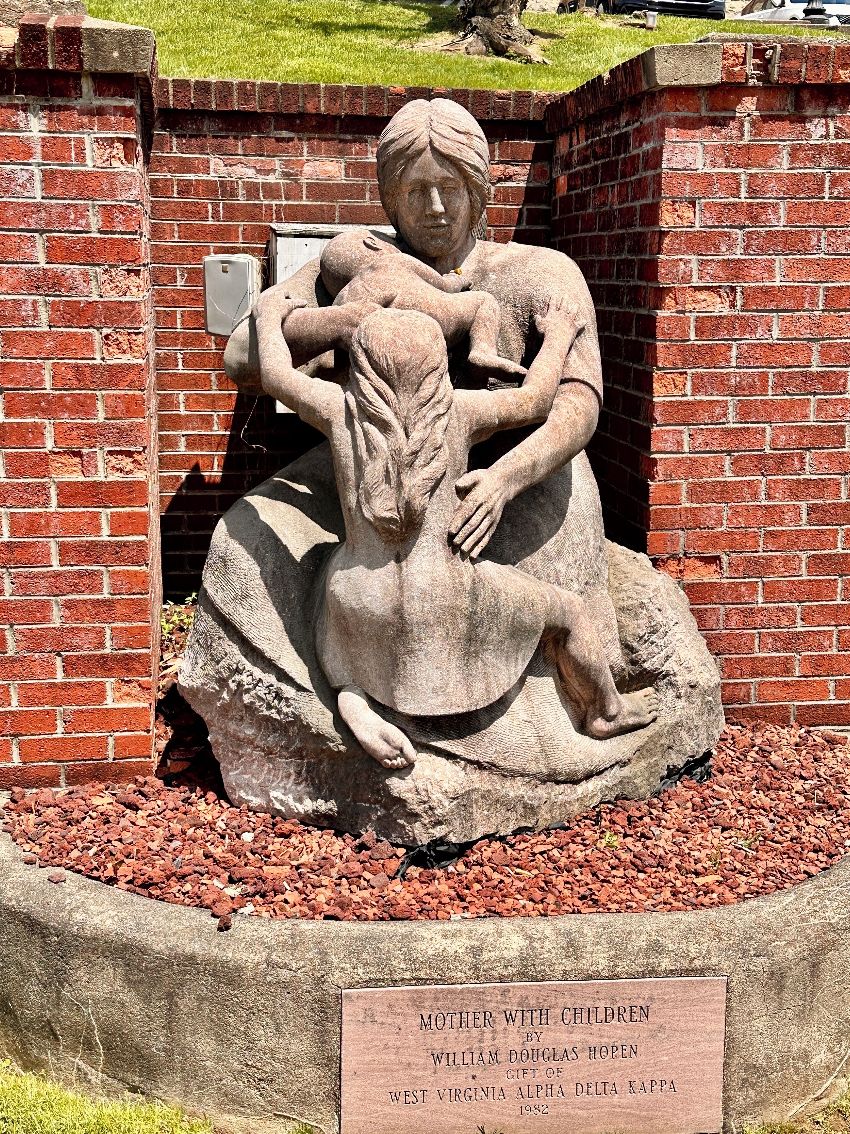 Una scultura in pietra intitolata 'Mother with Children' di William Douglas Hopen, situata in un'area paesaggistica con un muro in mattoni e uno sfondo erboso.