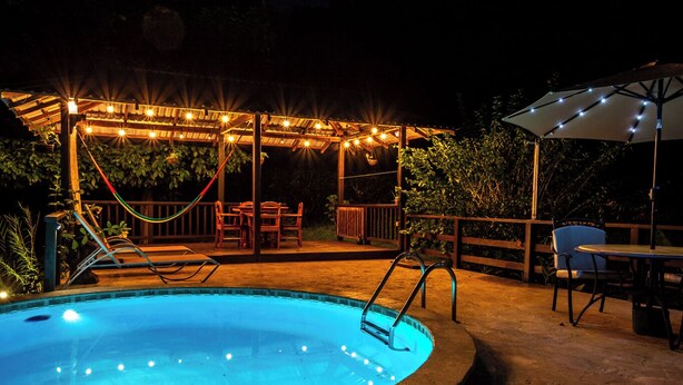 Une piscine avec une terrasse en bois et des guirlandes lumineuses, entourée de chaises et de tables.