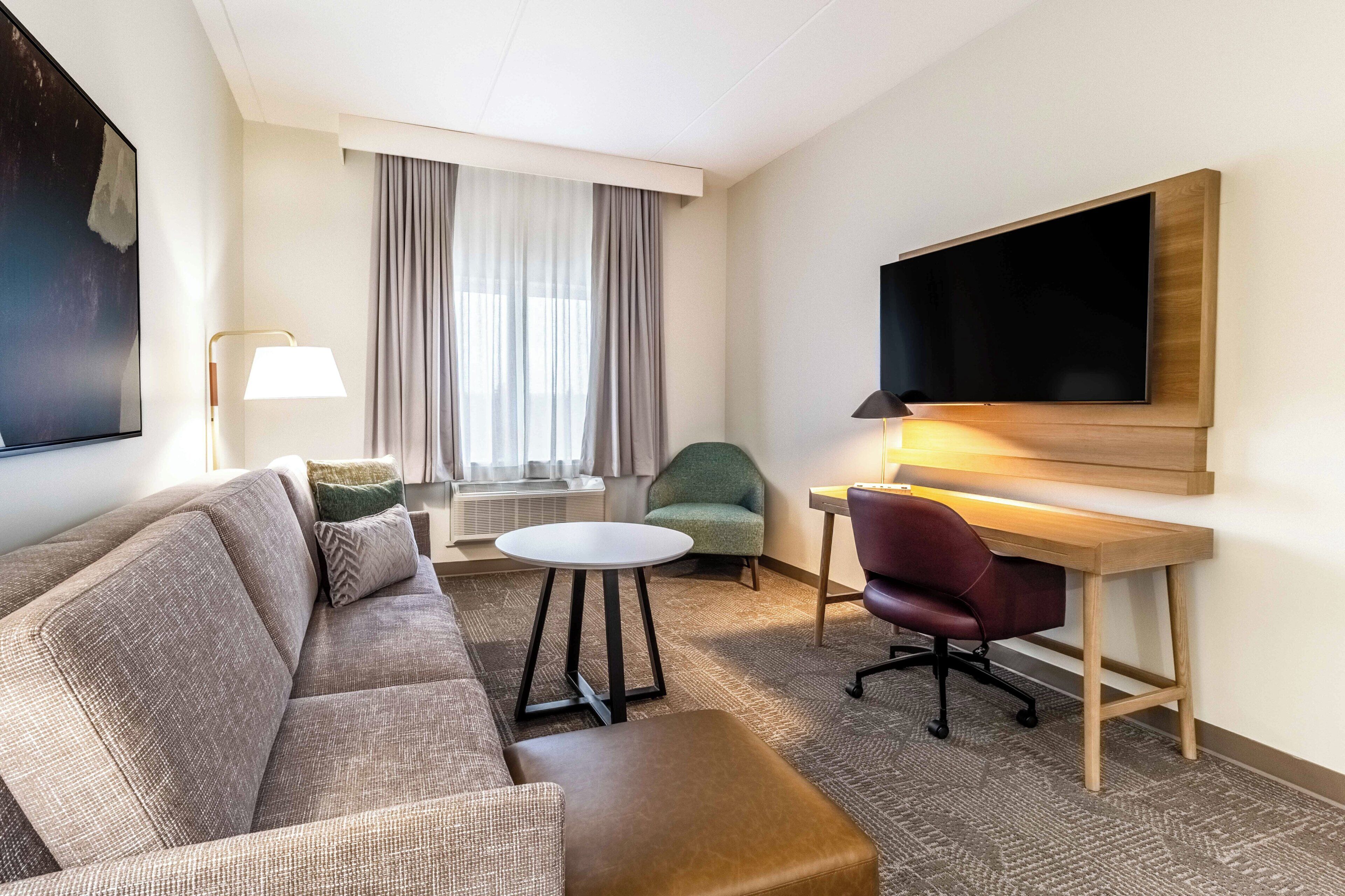 Habitación de hotel con sofá, una pequeña mesa redonda, escritorio con silla, televisor montado en la pared y ventana con cortinas.