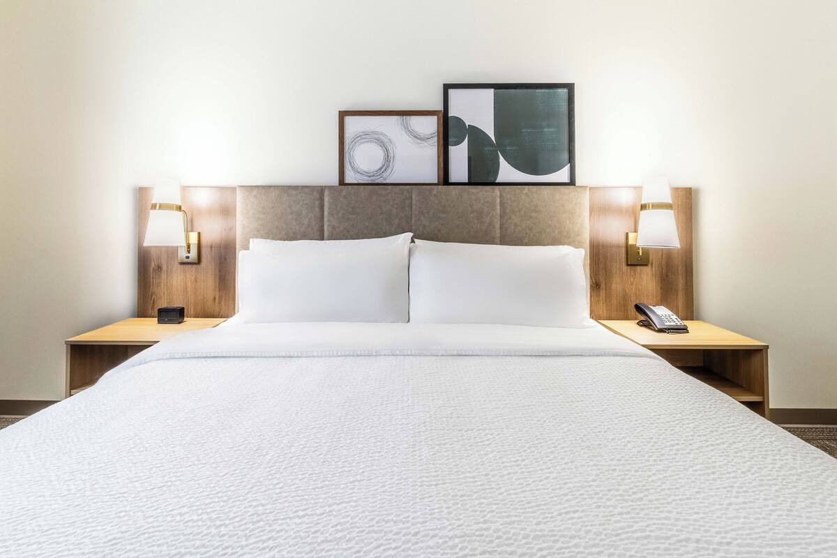 Una cama bien hecha con sábanas blancas, dos lámparas de noche, una obra de arte abstracta enmarcada y un teléfono en la mesita de noche.