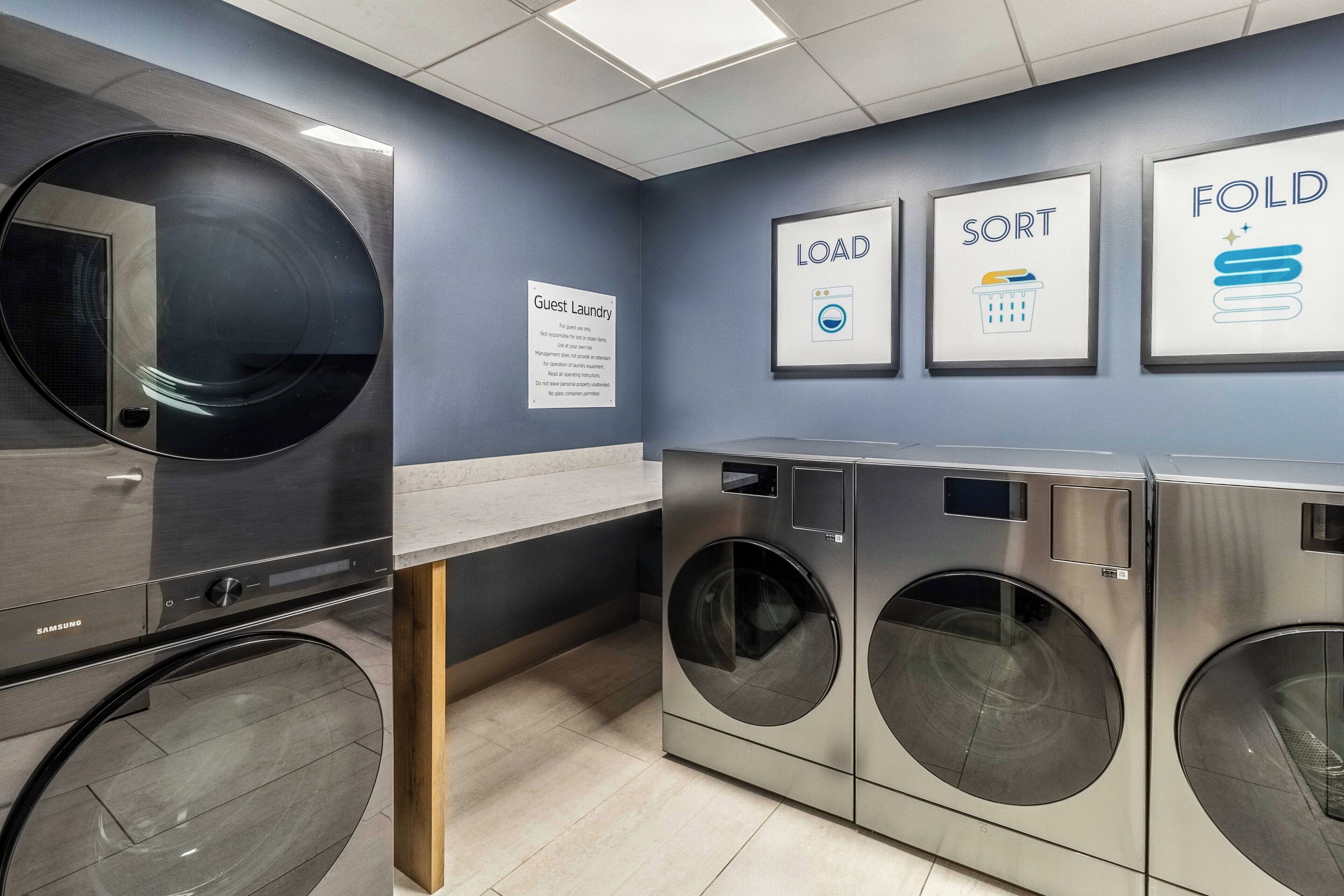 A modern laundry facility with a Samsung washer and dryer, a guest laundry sign, and wall-mounted signs indicating 'LOAD', 'SORT', and 'FOLD' instructions.
