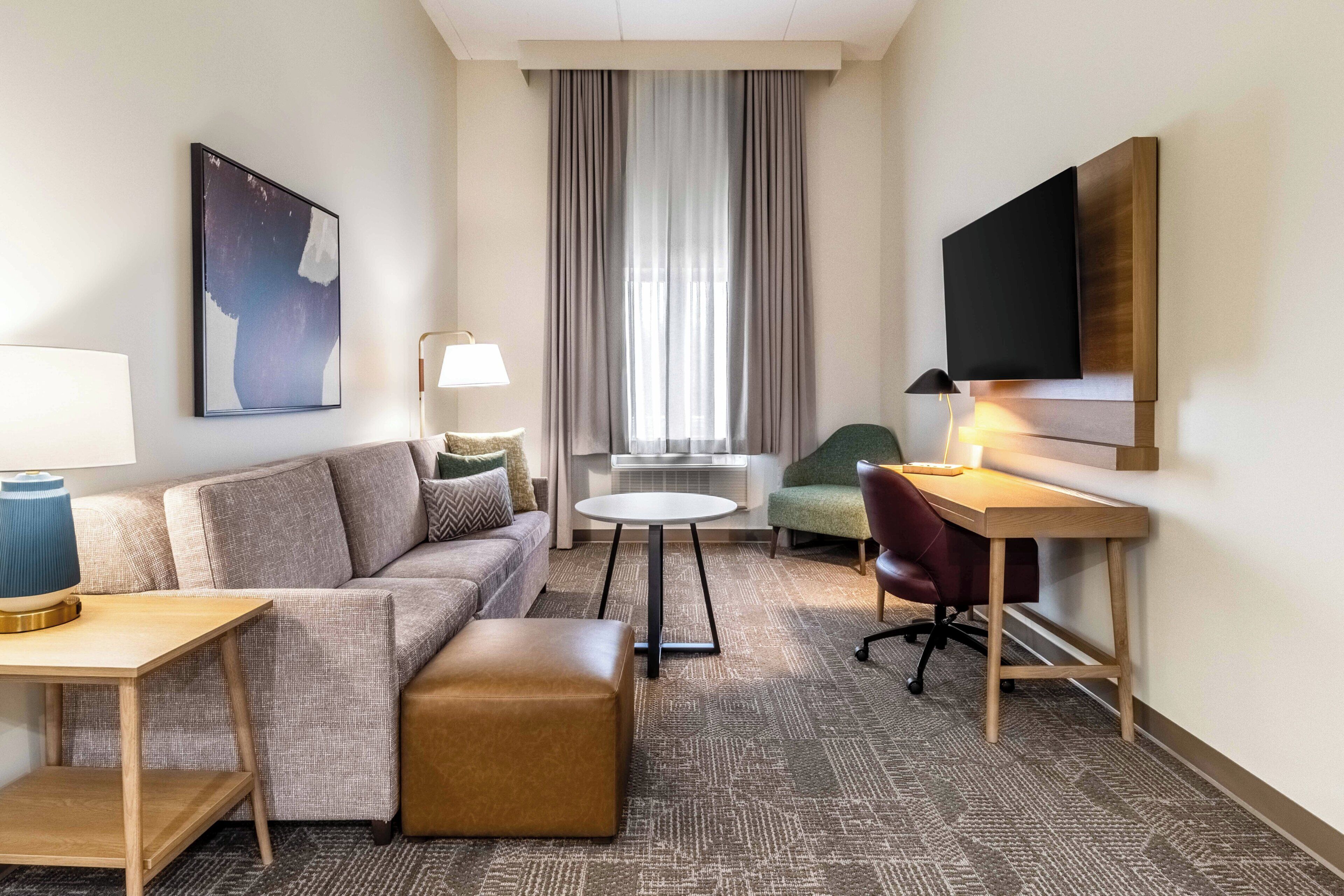 Hotelkamer met een bank, voetenbankje, klein rond tafeltje, bureau met stoel, televisie aan de muur en groot raam met gordijnen.