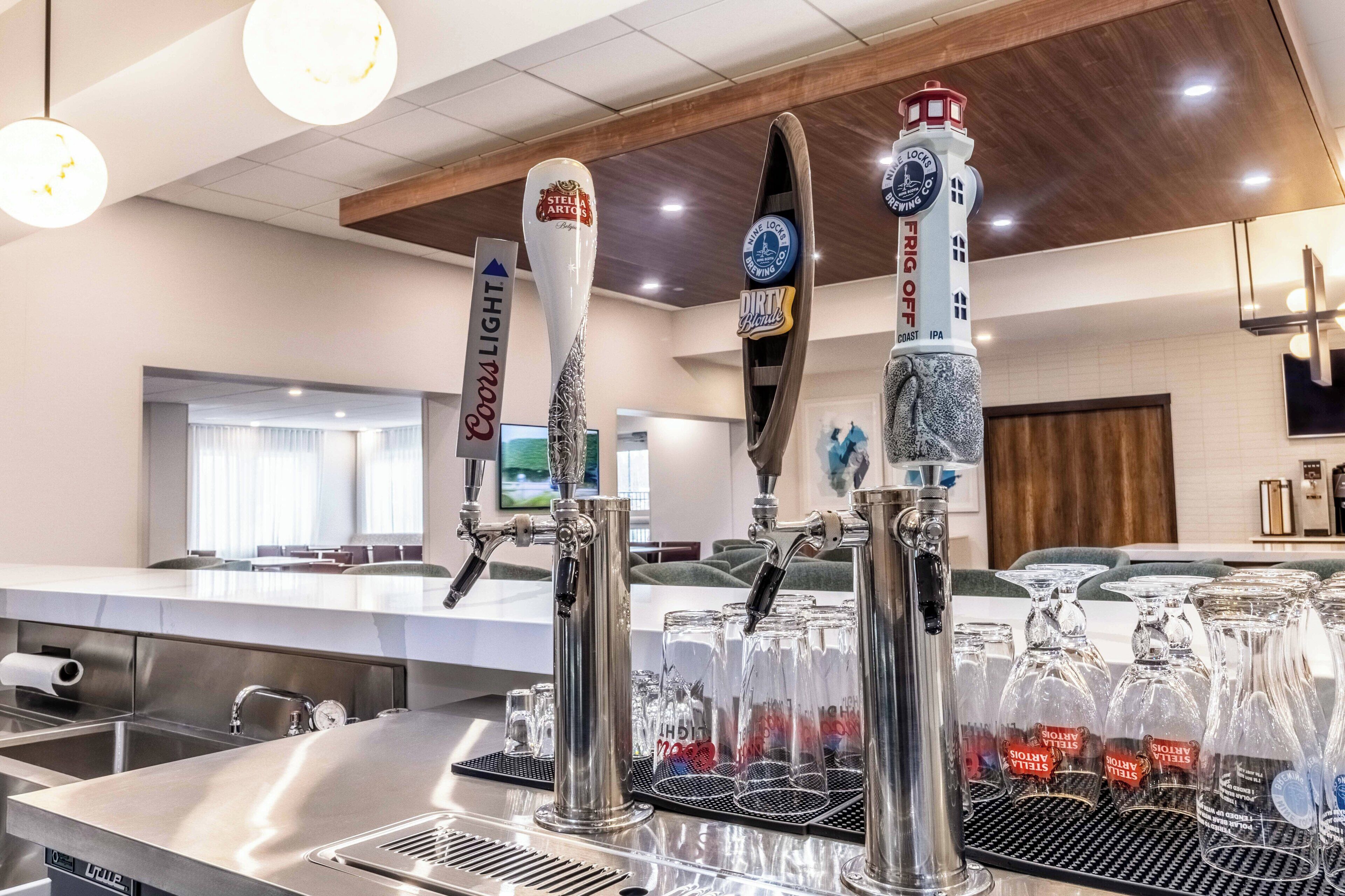 Una zona de bar con múltiples grifos identificados con marcas y tipos de cerveza, una barra con taburetes y vistas al salón de fondo.