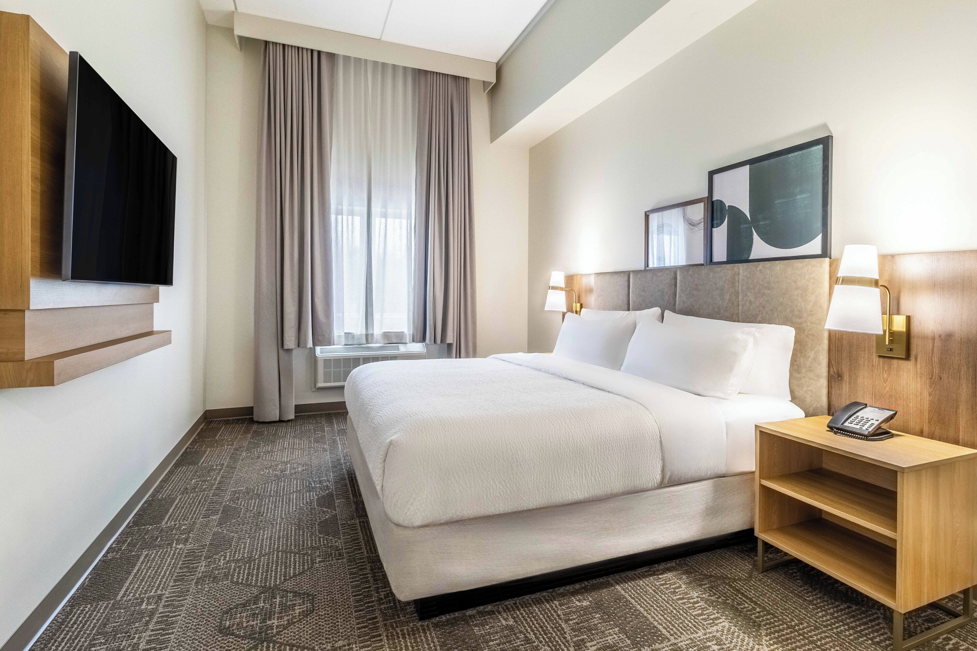 A hotel room with a large bed, a flat-screen TV mounted on the wall, a bedside table with a lamp, and a window with curtains.