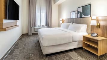 A hotel room with a large bed, a flat-screen TV mounted on the wall, a bedside table with a lamp, and a window with curtains.