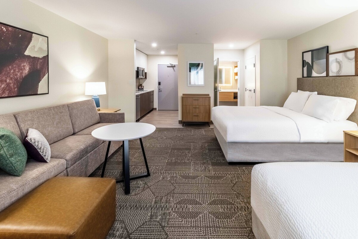 A hotel room with a sofa, a small round table, a bed, a kitchenette, and a bathroom.