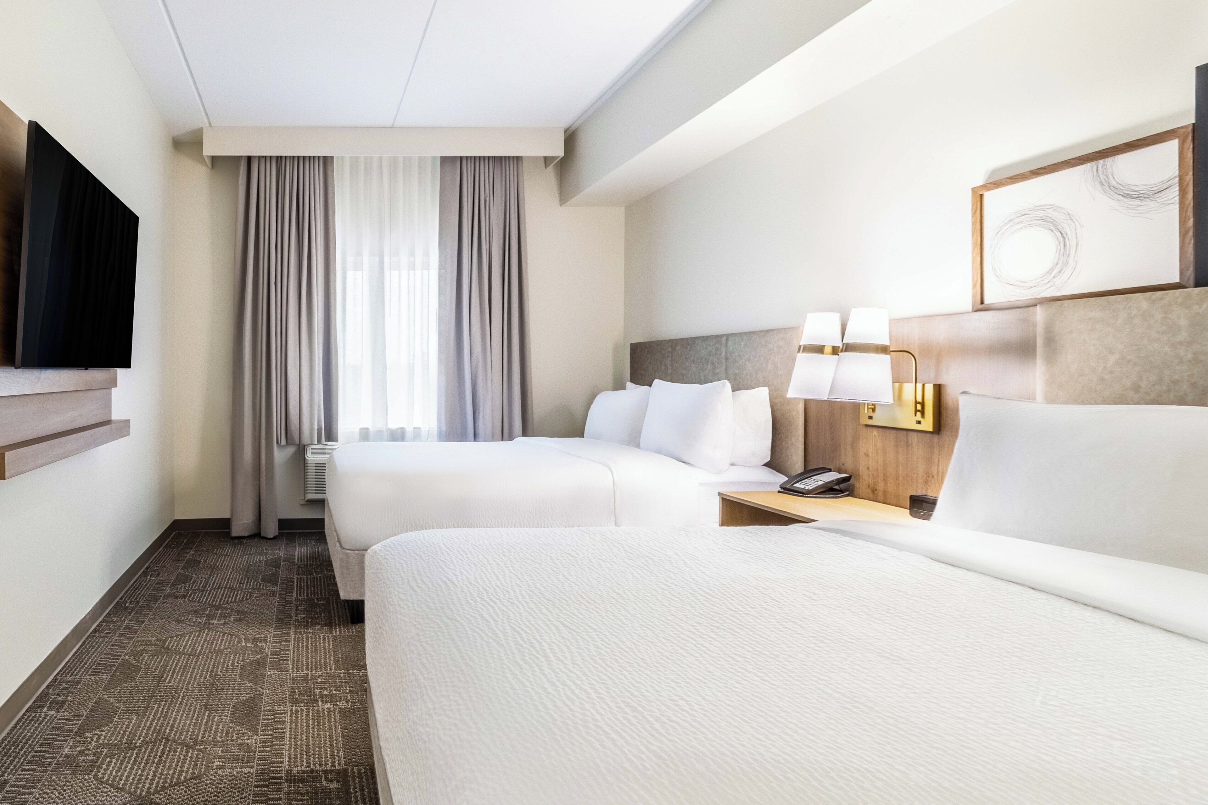 Une chambre d’hôtel avec deux lits, un téléviseur à écran plat fixé au mur, une table de chevet avec des lampes et une grande fenêtre avec des rideaux.