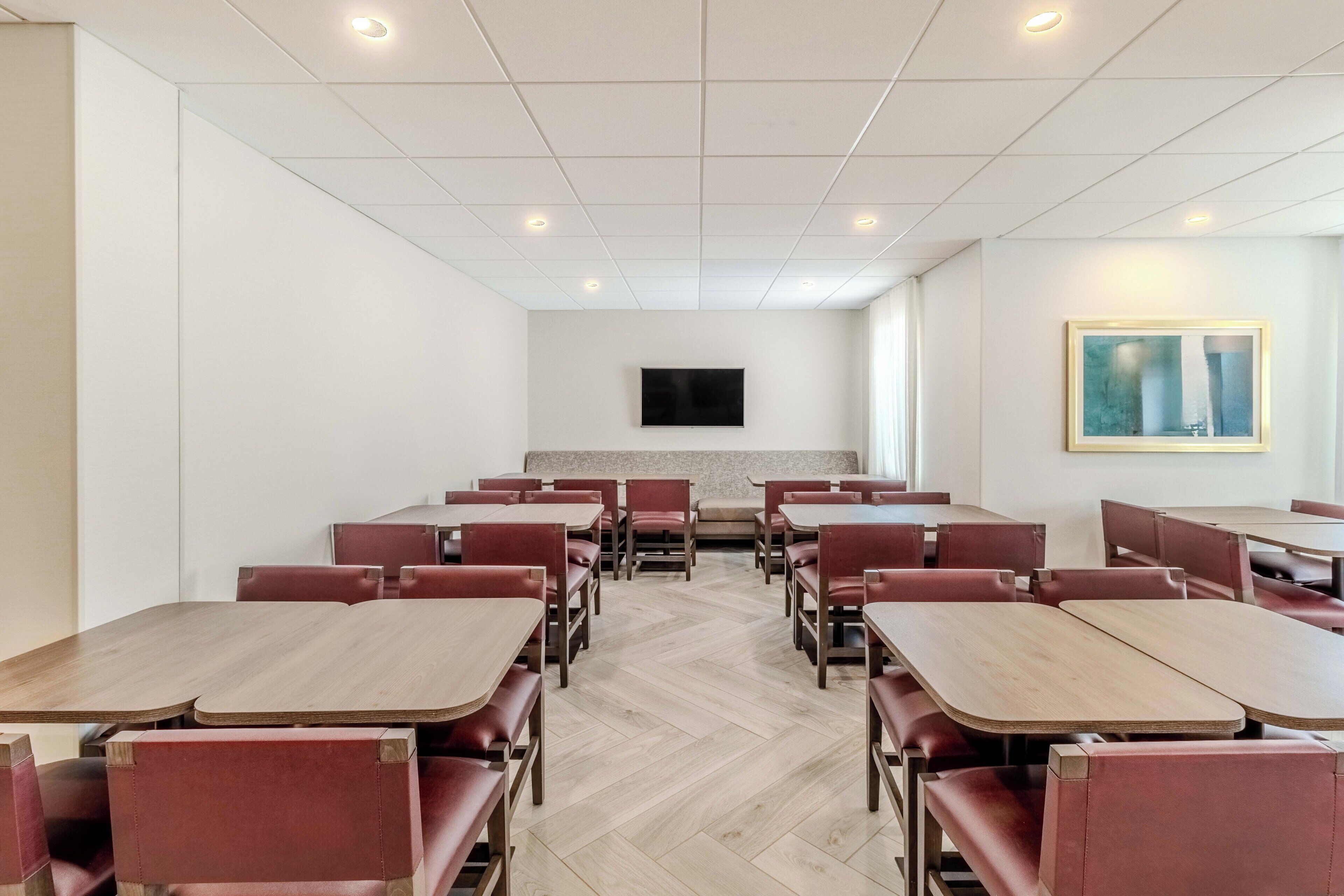 A dining area with multiple tables and chairs, a flat-screen TV mounted on the wall, and a herringbone-patterned floor.