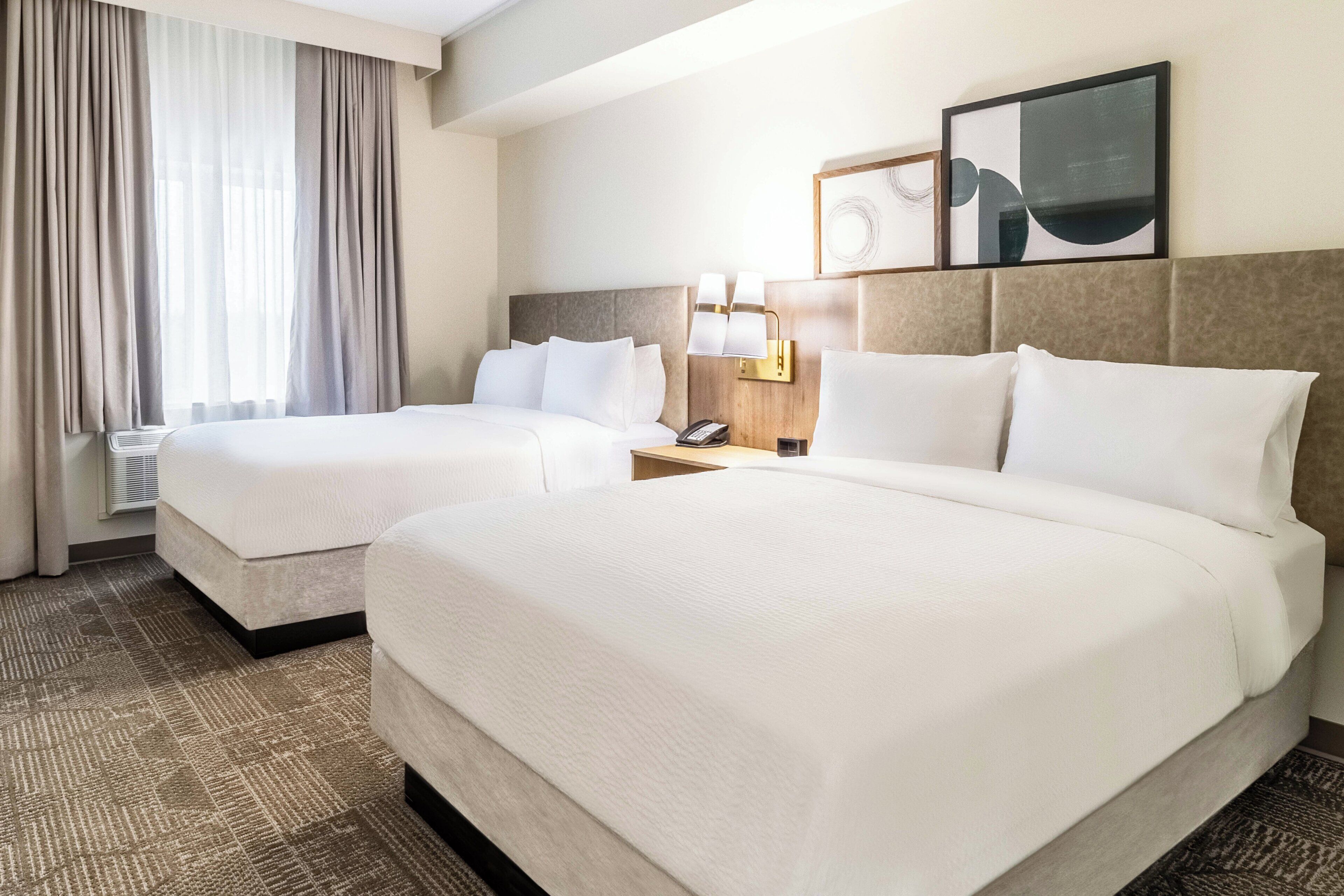 A hotel room with two beds, a large window with curtains, a bedside table with a lamp, and a framed picture on the wall.