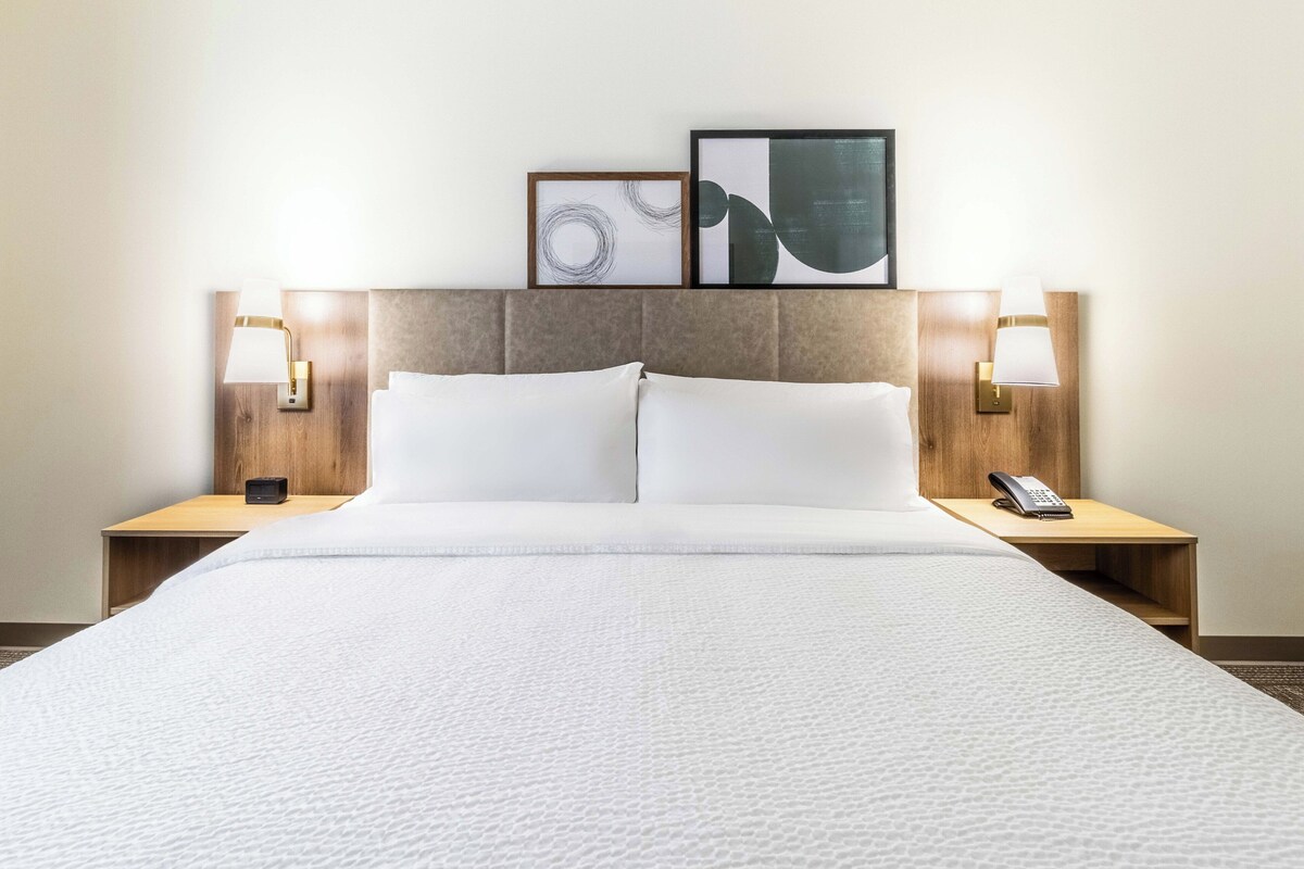 A neatly made bed with white linens, two bedside lamps, a framed abstract artwork, and a phone on the nightstand.
