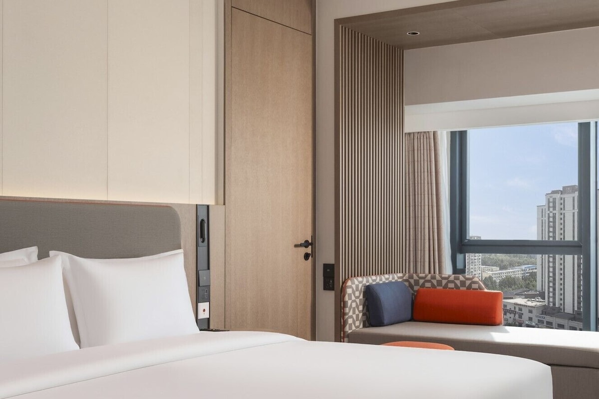 A hotel room with a bed, a seating area with cushions, a window with a city view, and a door.