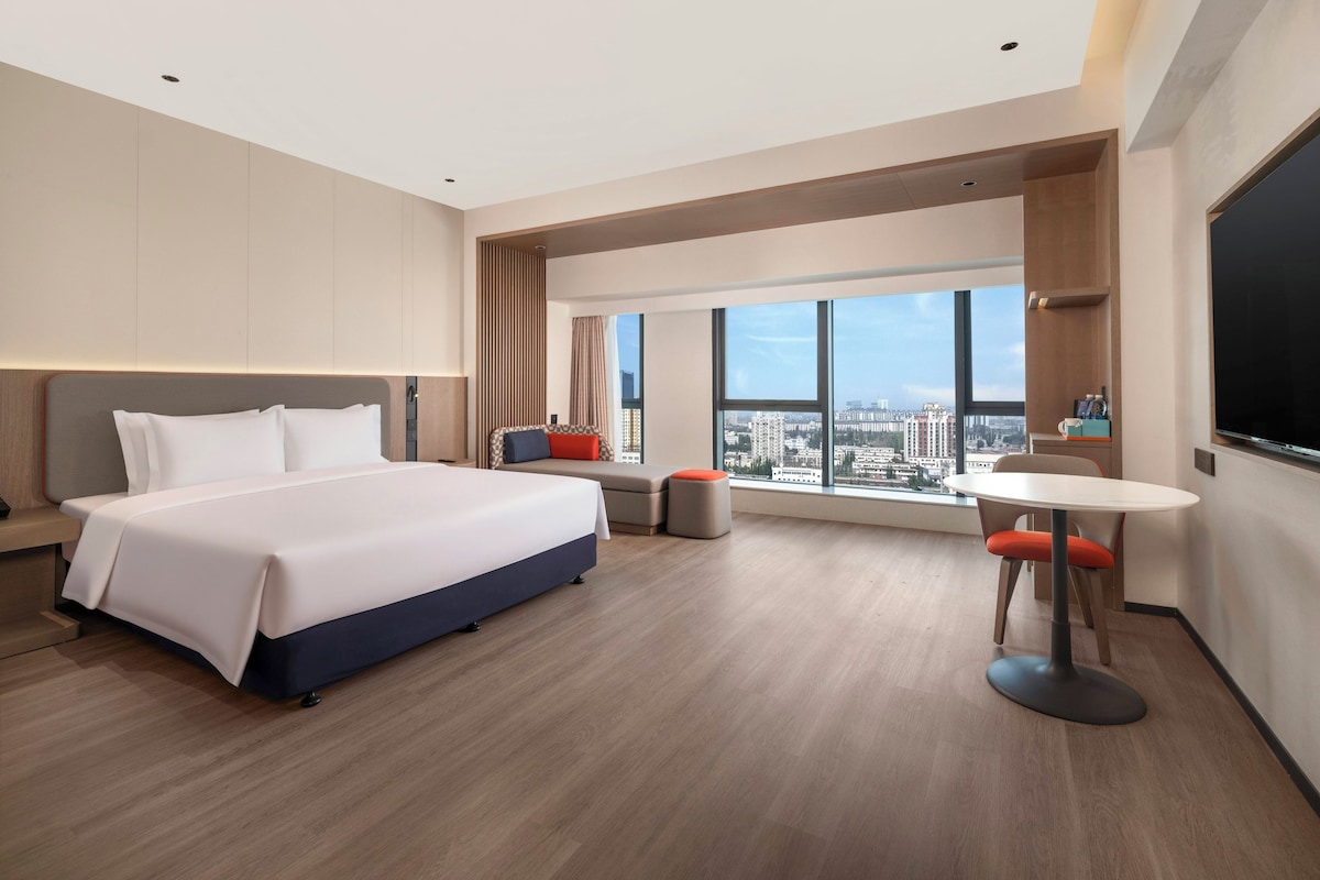 A modern hotel room with a large bed, a small seating area, a dining table, and a city view through large windows.