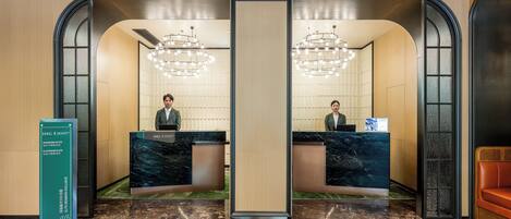 A hotel reception area with two receptionists, a marble reception desk, a chandelier, and a sign indicating 'Diamond Elite' and 'Platinum Elite' membership levels.
