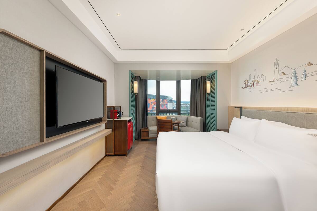 A hotel room with a large bed, a flat-screen TV, a desk with a red coffee machine, a sofa, and a city view through the window.