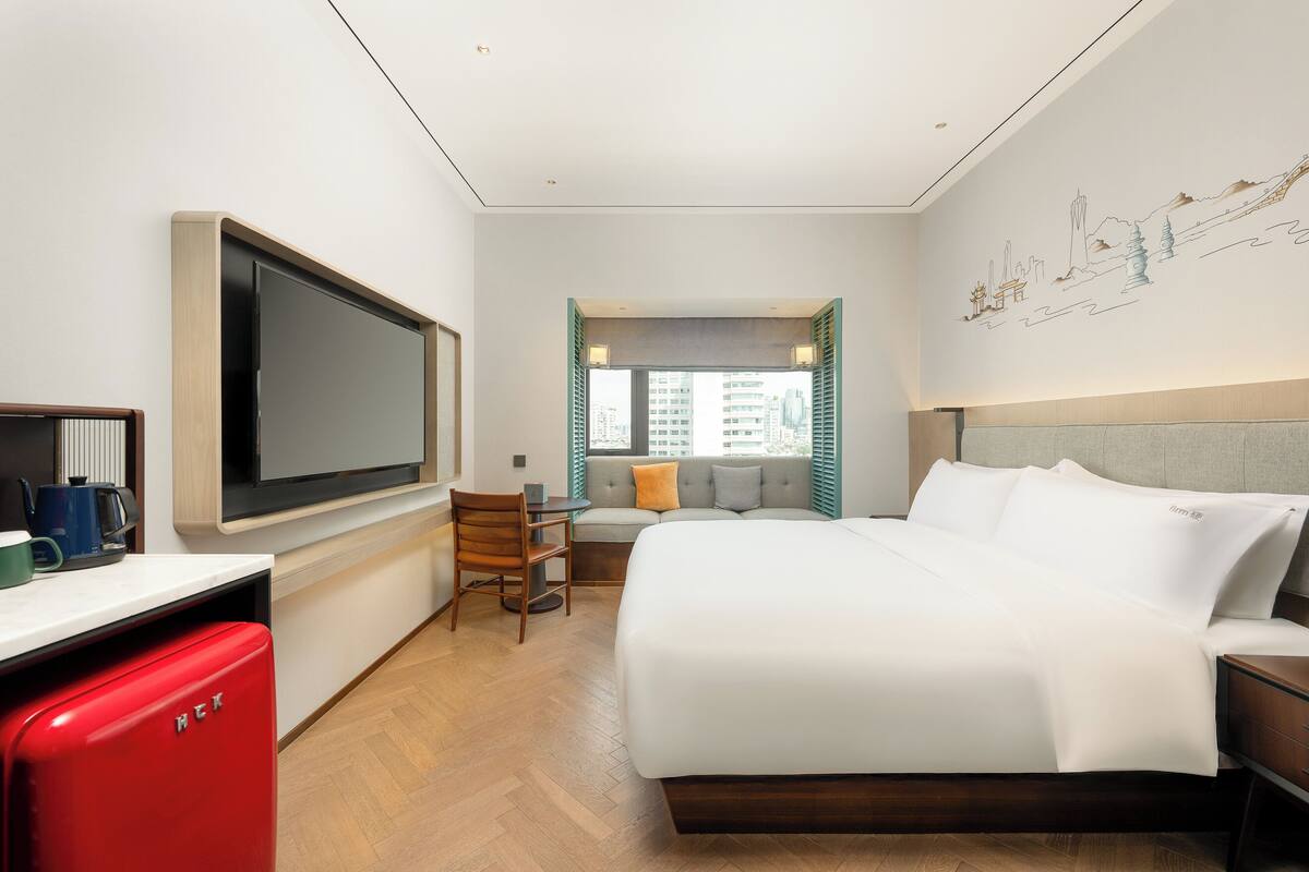 A modern hotel room with a large bed, a TV, a desk with a red appliance, and a window with a city view.