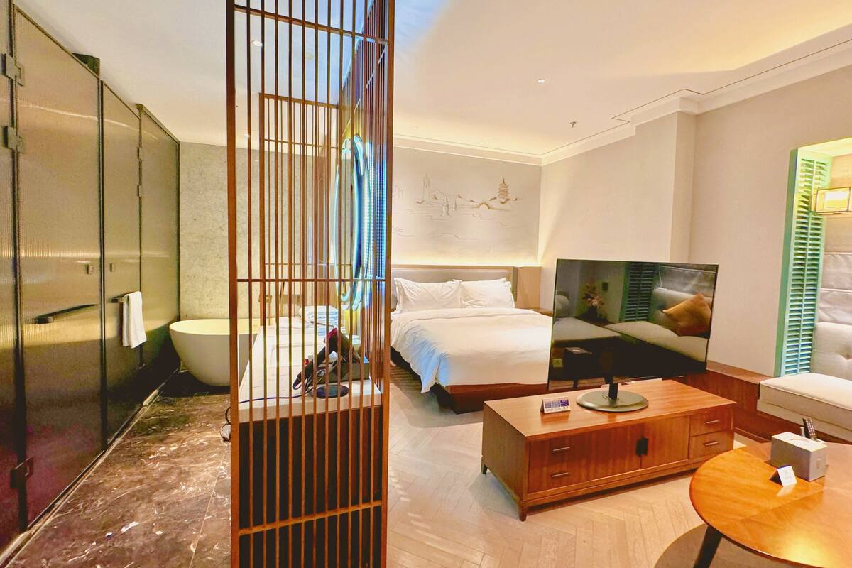 A hotel room with a bed, a wooden cabinet, a round table, a television, and a glass shower enclosure.