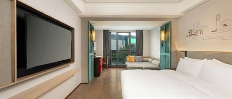A hotel room with a large bed, a flat-screen TV mounted on the wall, a seating area with a sofa and chairs, and a view through the window.