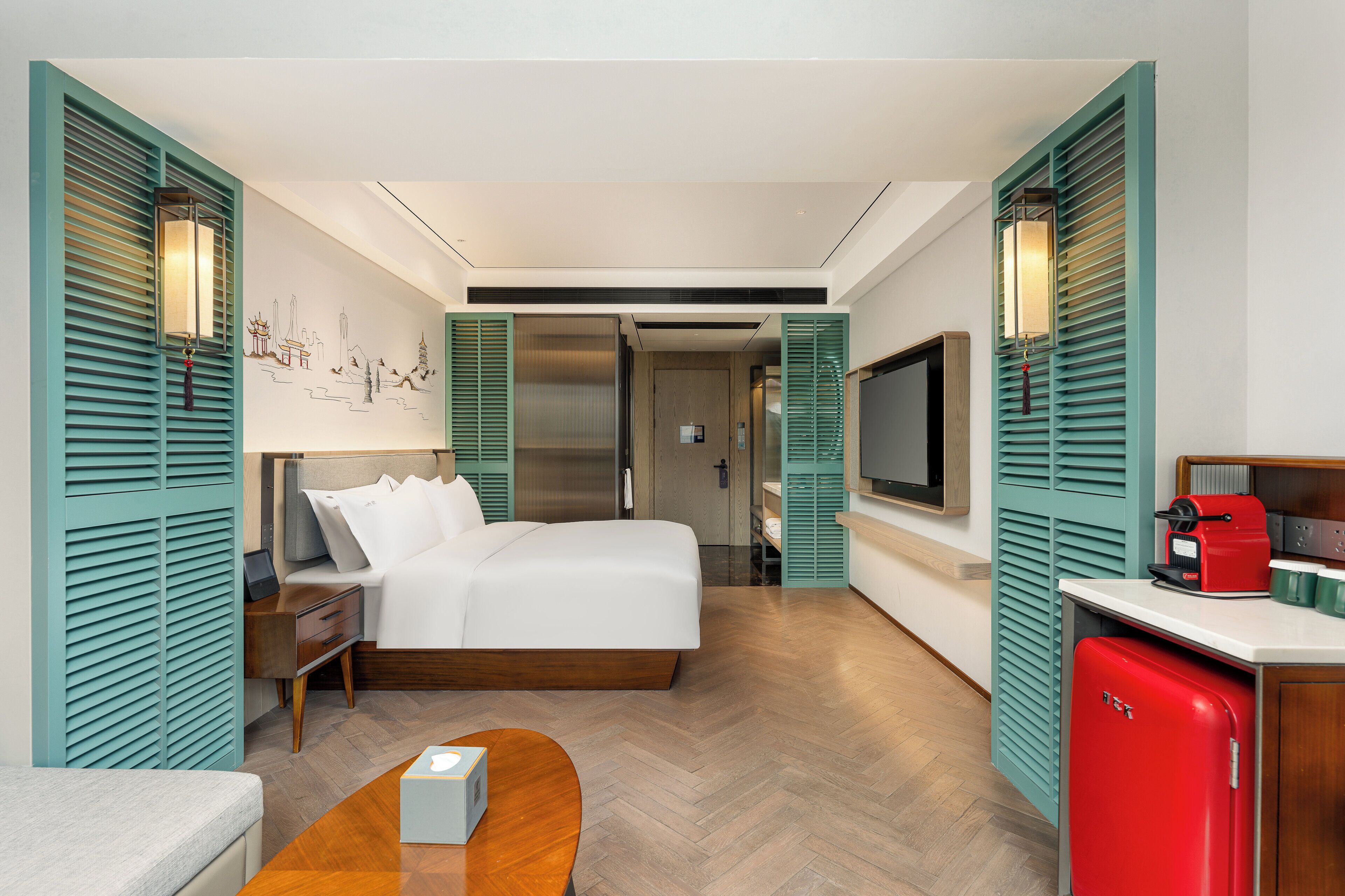 A modern hotel room with a bed, bedside table, lamp, TV, and a small red refrigerator.