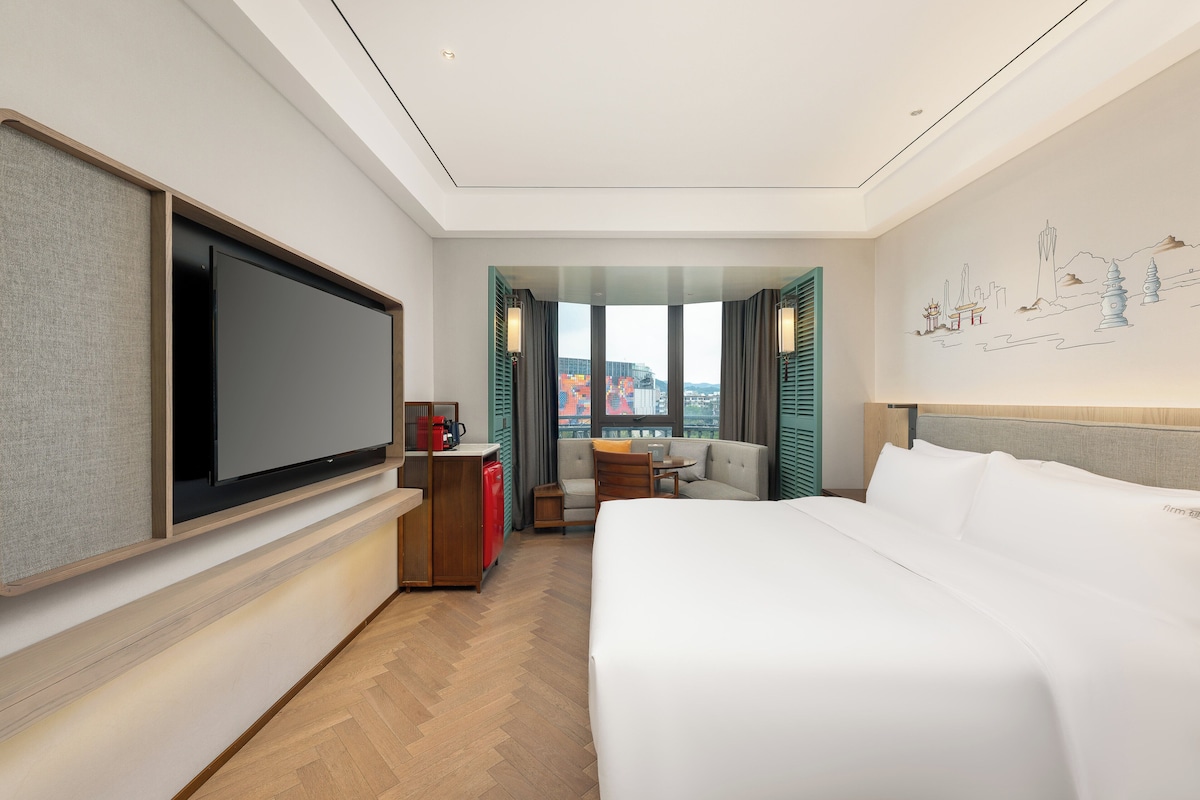 Habitación de hotel con una cama grande, televisor de pantalla plana, escritorio con cafetera roja, sofá y vista a la ciudad desde la ventana.