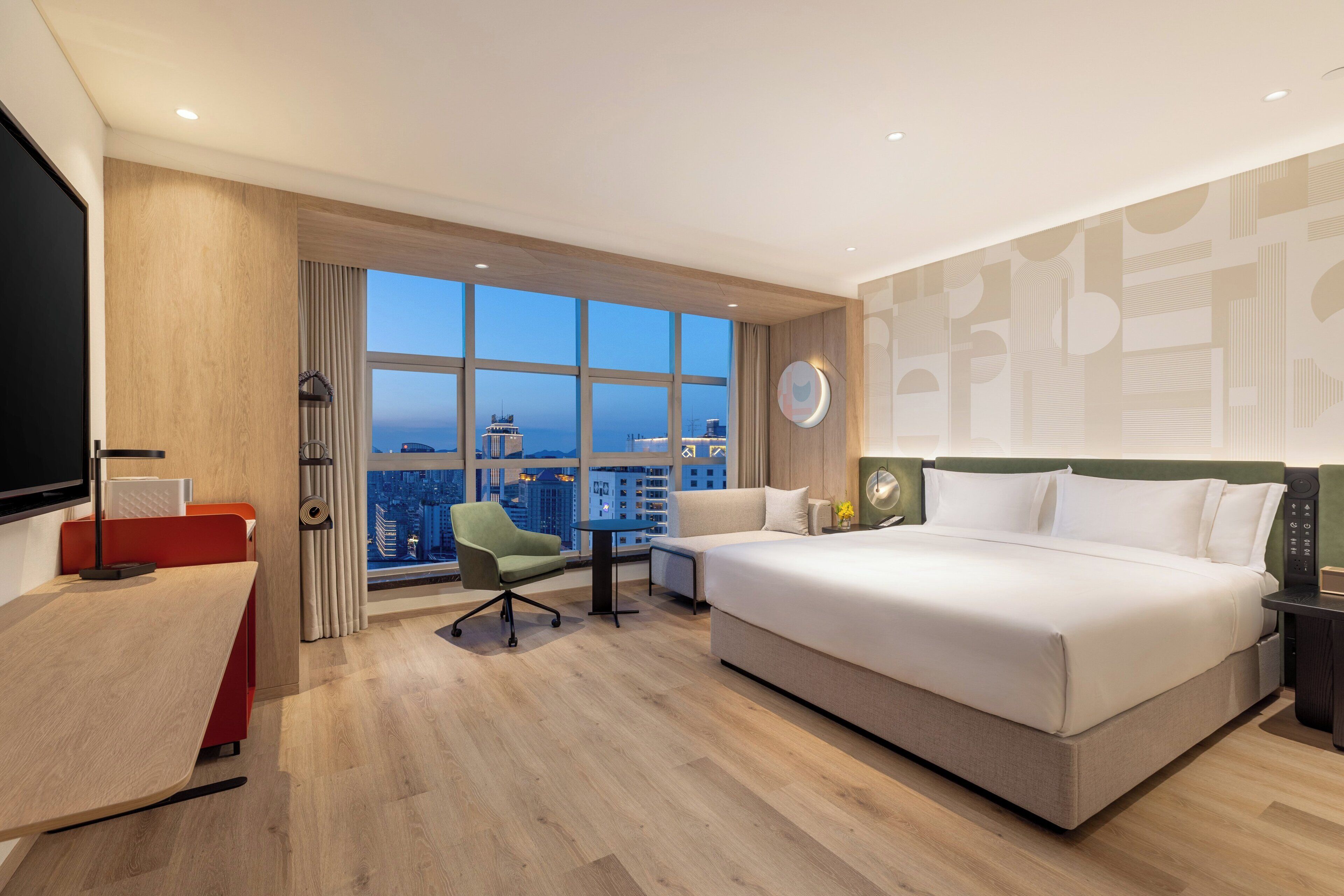 A hotel room with a large bed, a desk, a chair, and a view of the cityscape.