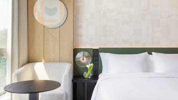A hotel room with a bed, a small table, a chair, a nightstand, a wall clock, and a vase with flowers.