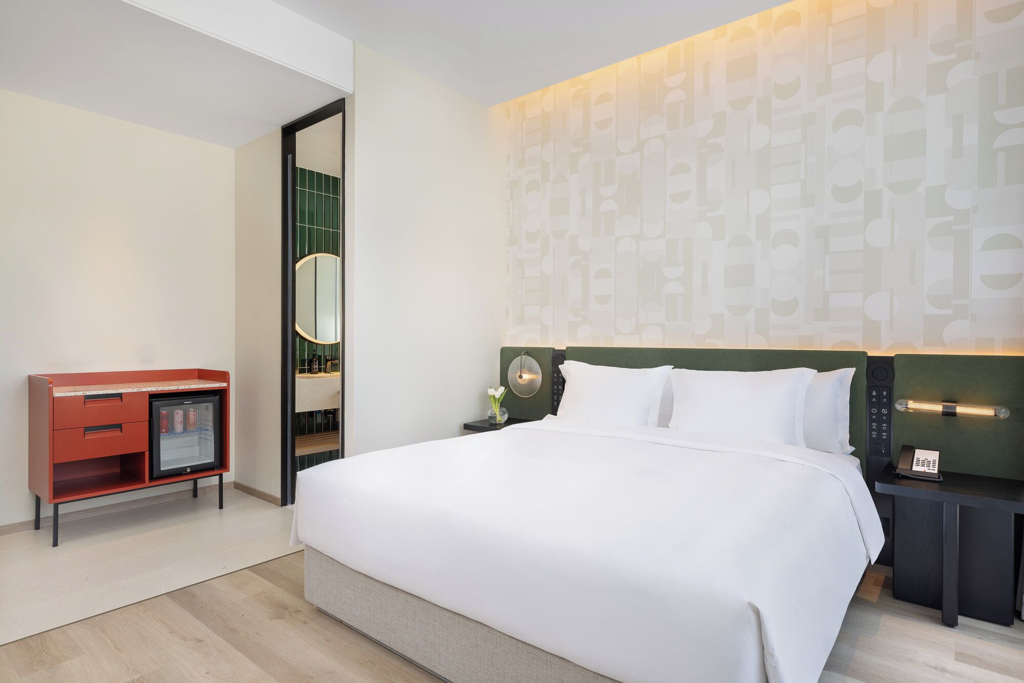 Quarto de hotel com uma cama grande, mesa de cabeceira com abajur, pequena escrivaninha com refrigerador e um espelho.