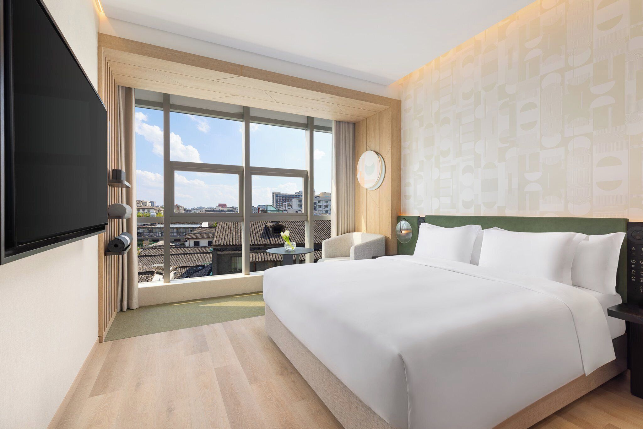 A hotel room with a large bed, a TV mounted on the wall, a round clock, and a view of the city through large windows.