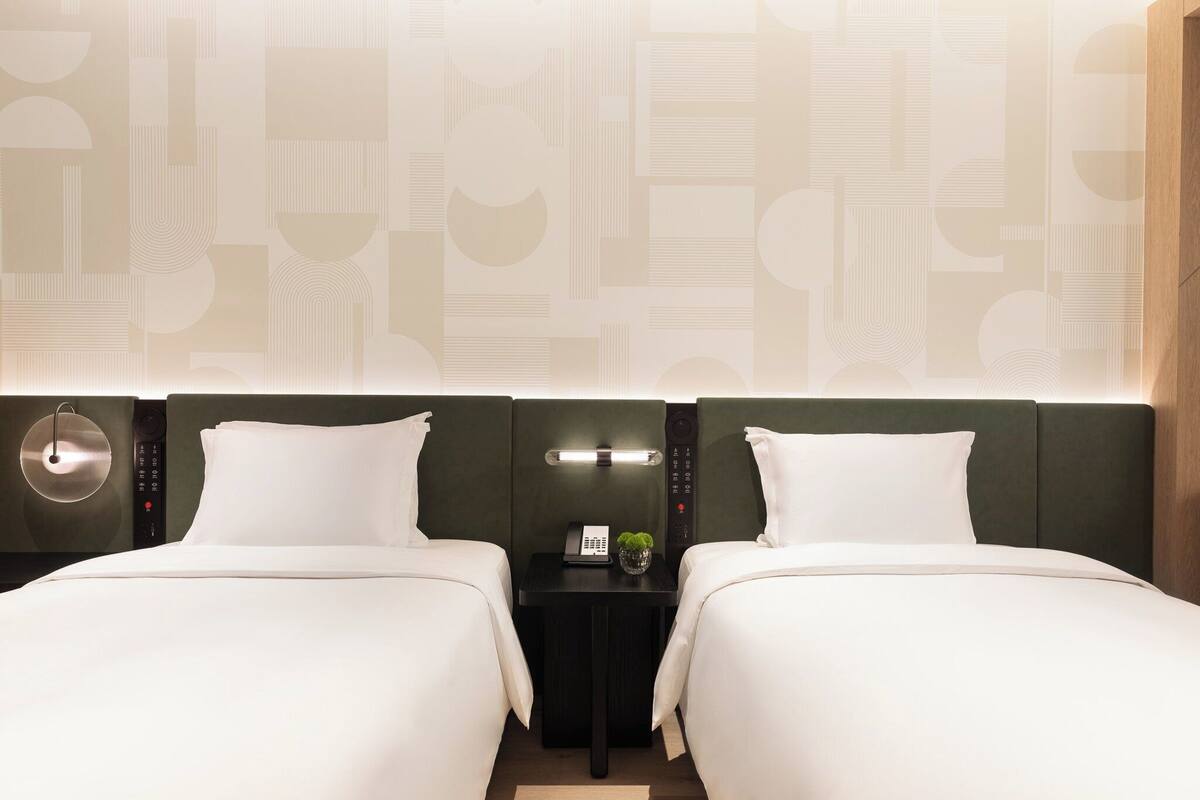Habitación de hotel con dos camas, una mesita de noche con lámpara, teléfono y una planta pequeña.