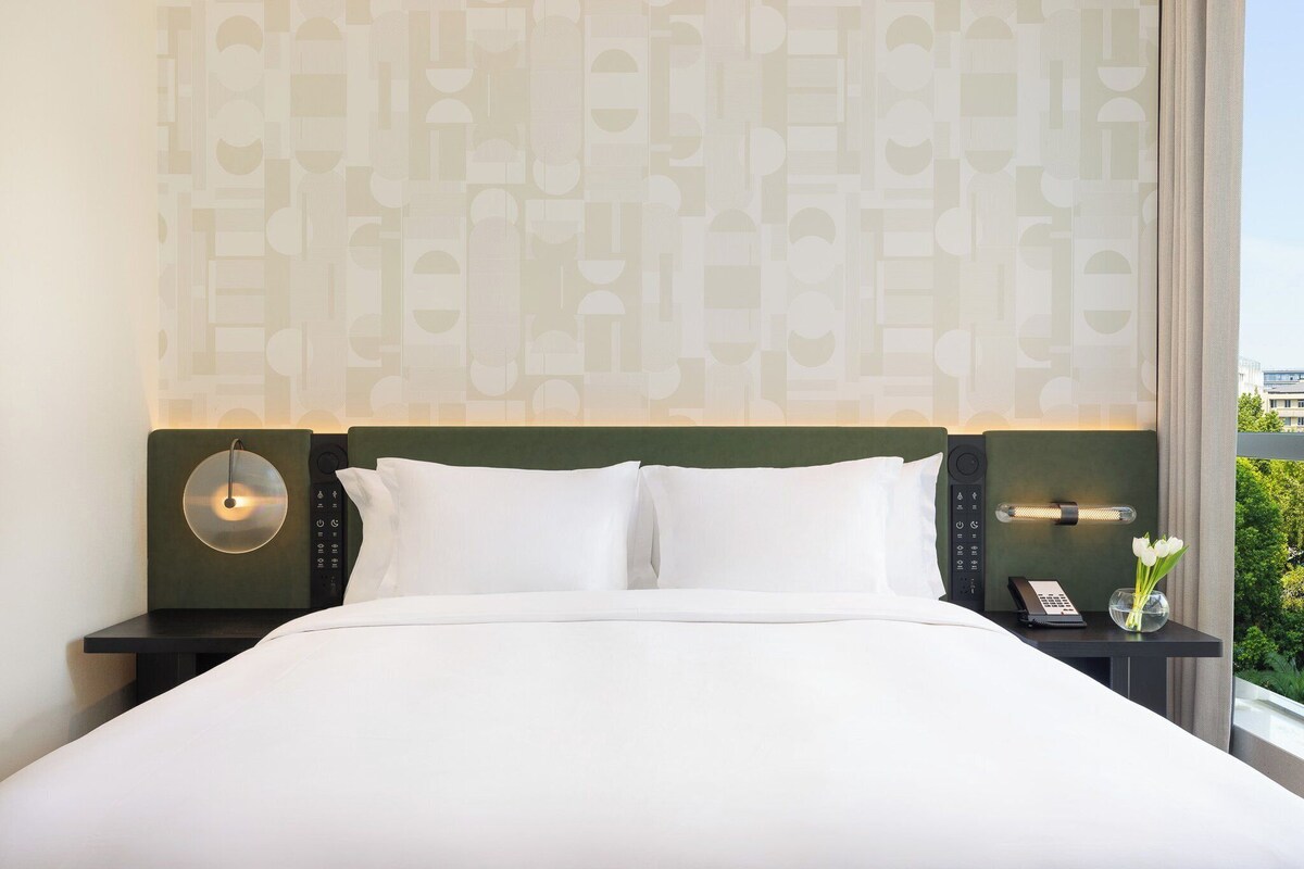 A neatly made bed with white linens, a bedside lamp, a phone, and a vase with flowers on a nightstand.