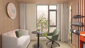 Habitación de hotel moderna con sofá, una silla verde, una pequeña mesa redonda, ventana con vistas a zonas verdes y una pared de madera con espejo y estanterías.