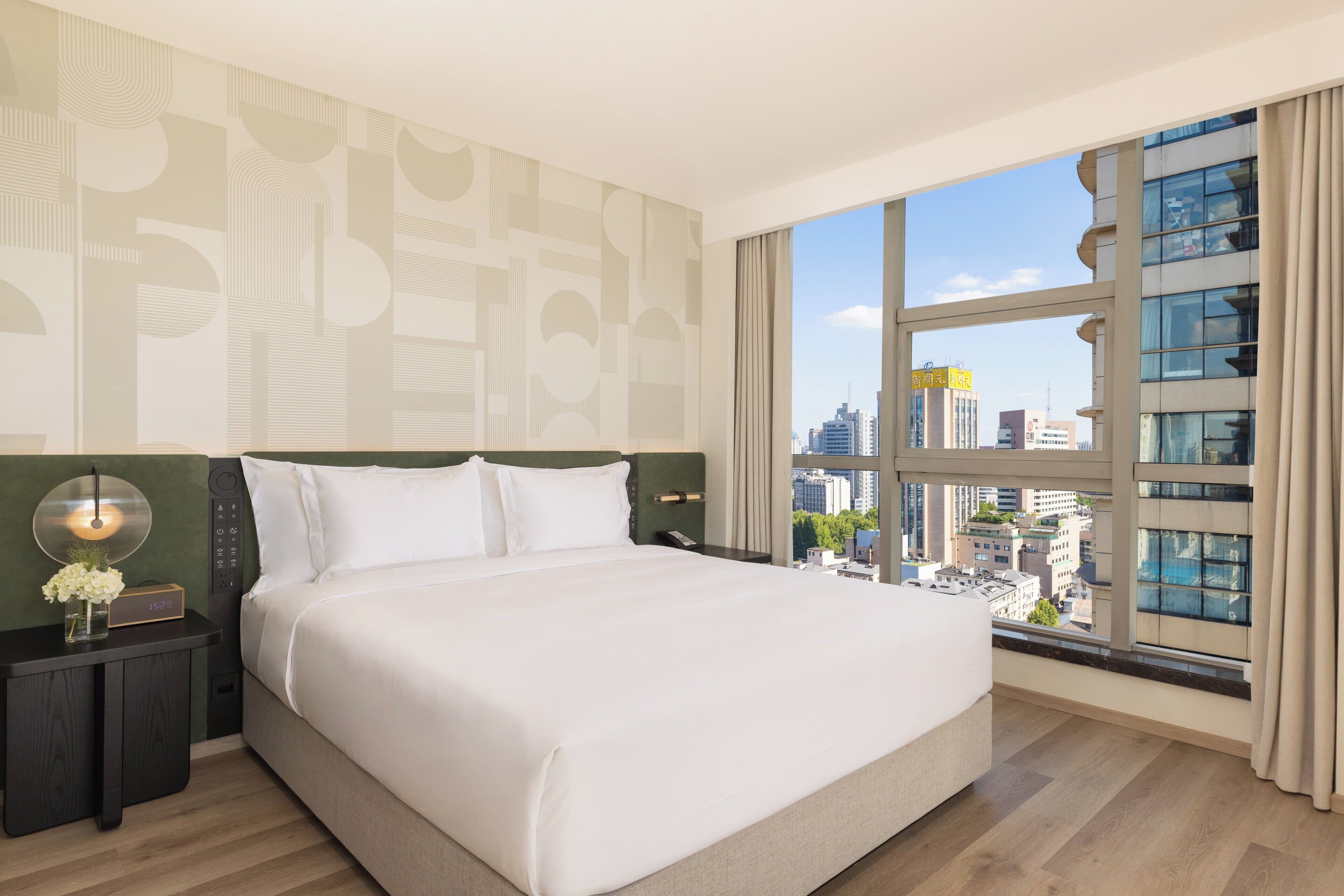 A hotel room with a large bed, a nightstand, a digital clock, and a view of the city through a large window.