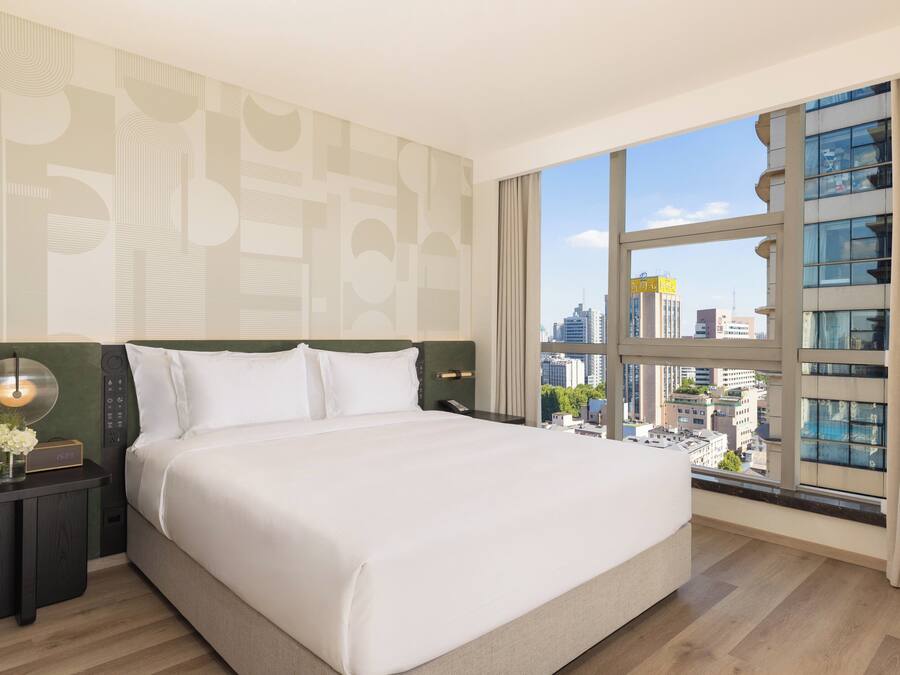 A hotel room with a large bed, a nightstand, a digital clock, and a view of the city through a large window.